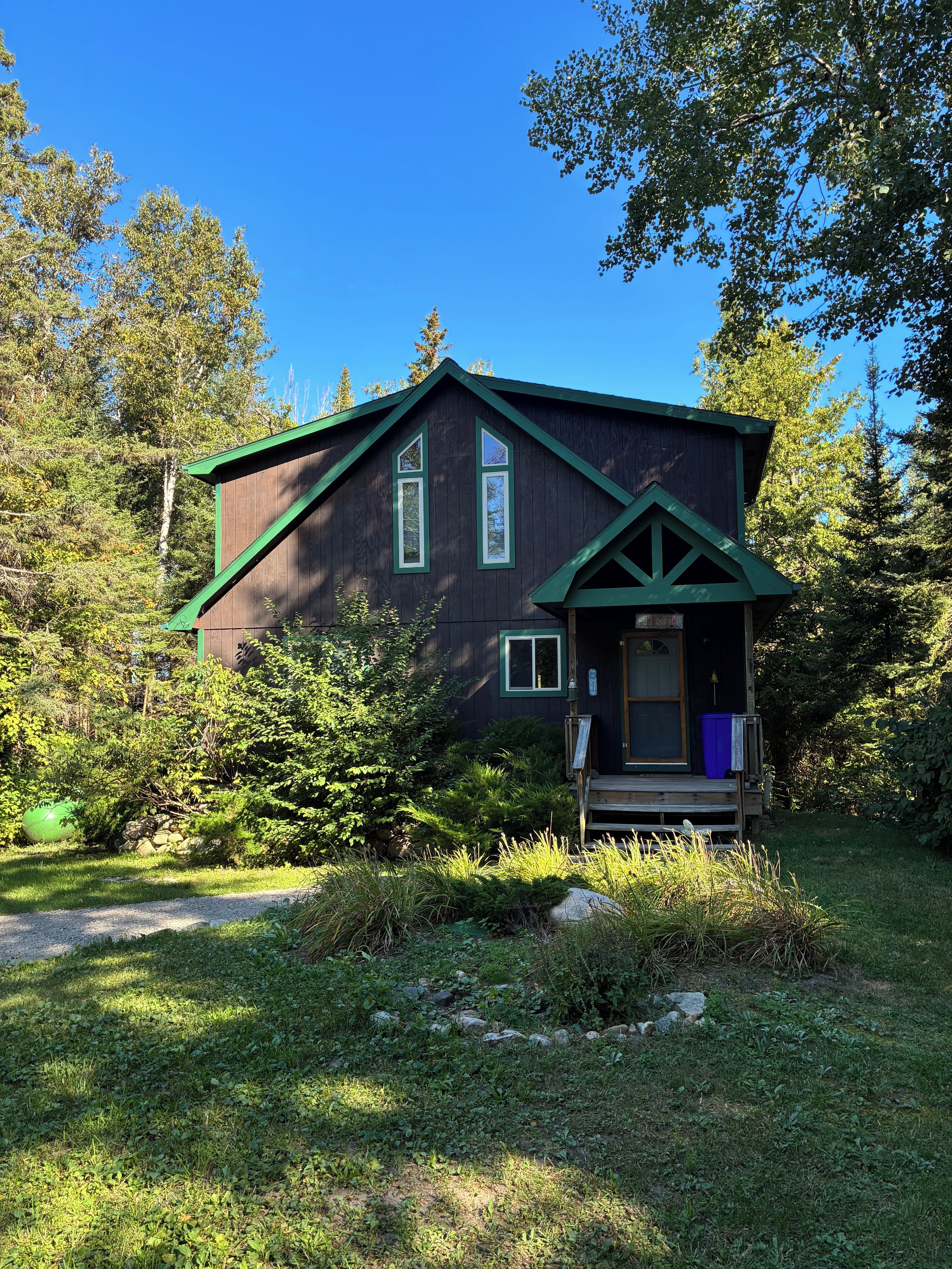 A cabin with a dark wooden exterior, green trim, and a porch with a blue trash bin.