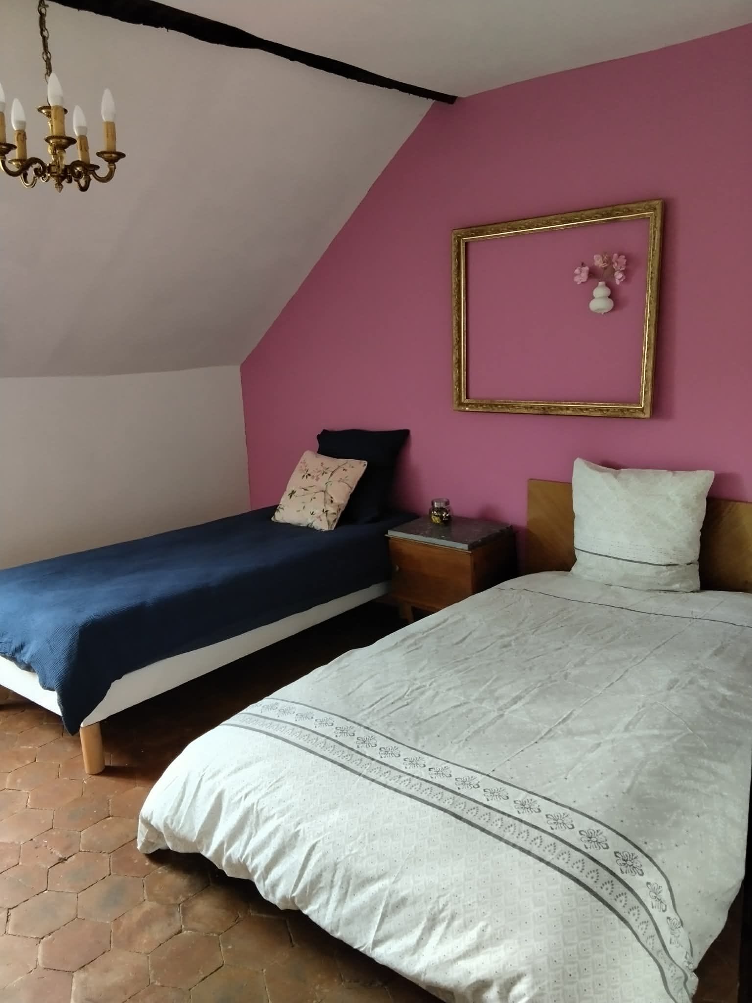 Une chambre avec deux lits, un lustre, un mur rose et un tableau encadré.
