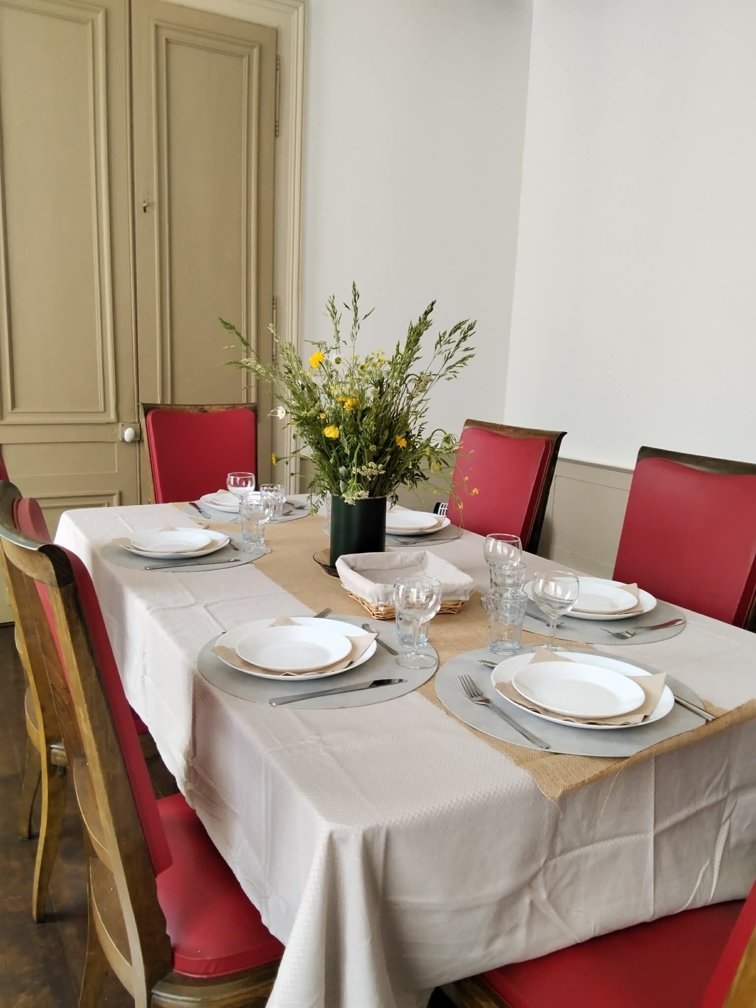 Mesa de jantar posta para quatro com toalhas brancas, cadeiras vermelhas e um vaso de flores.