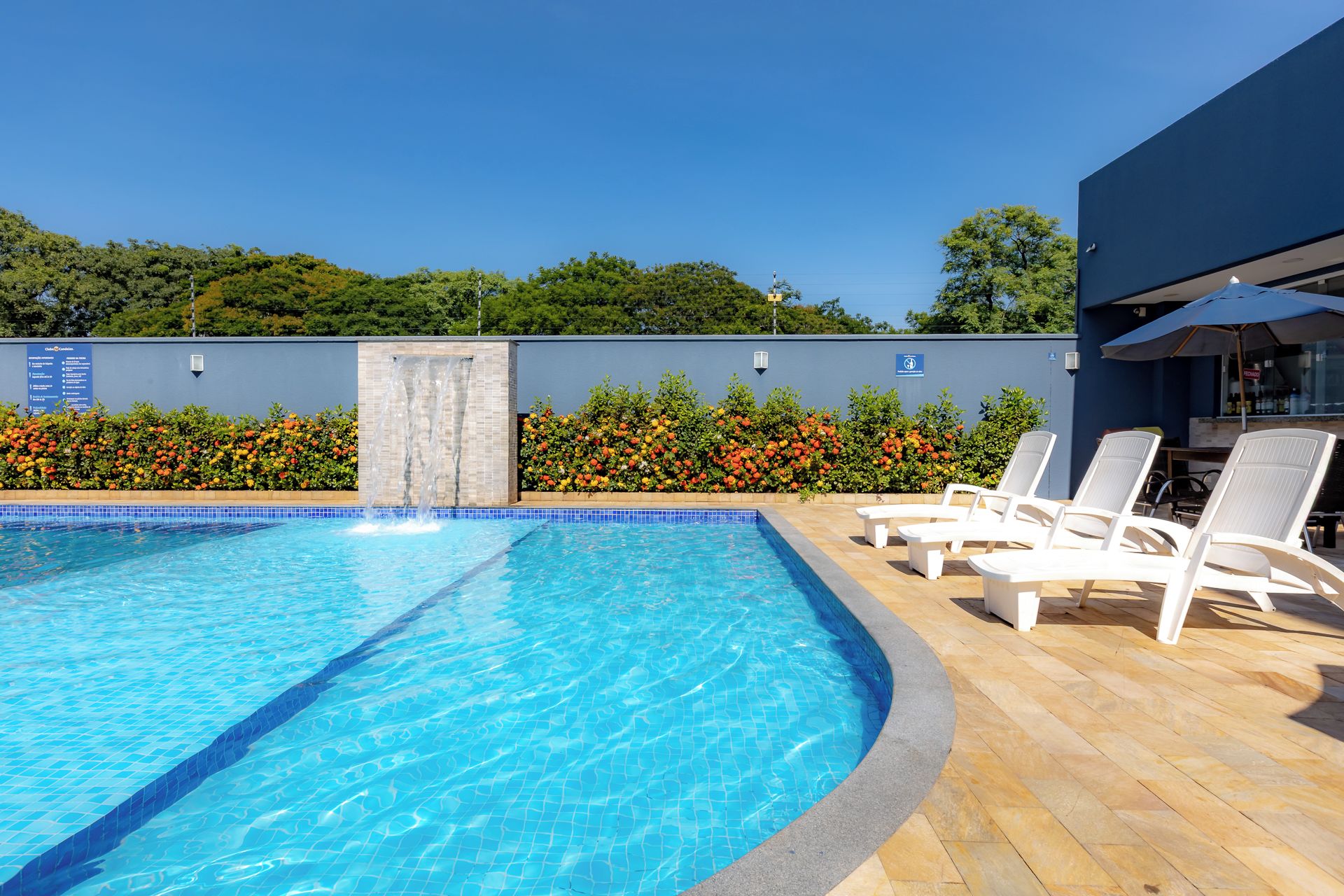 Una piscina con bordo piastrellato, circondata da un pontile in legno e un’area relax con poltrone prendisole.