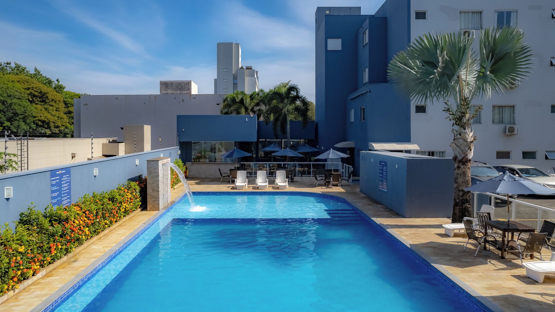 A swimming pool with lounge chairs and umbrellas, surrounded by buildings and palm trees.