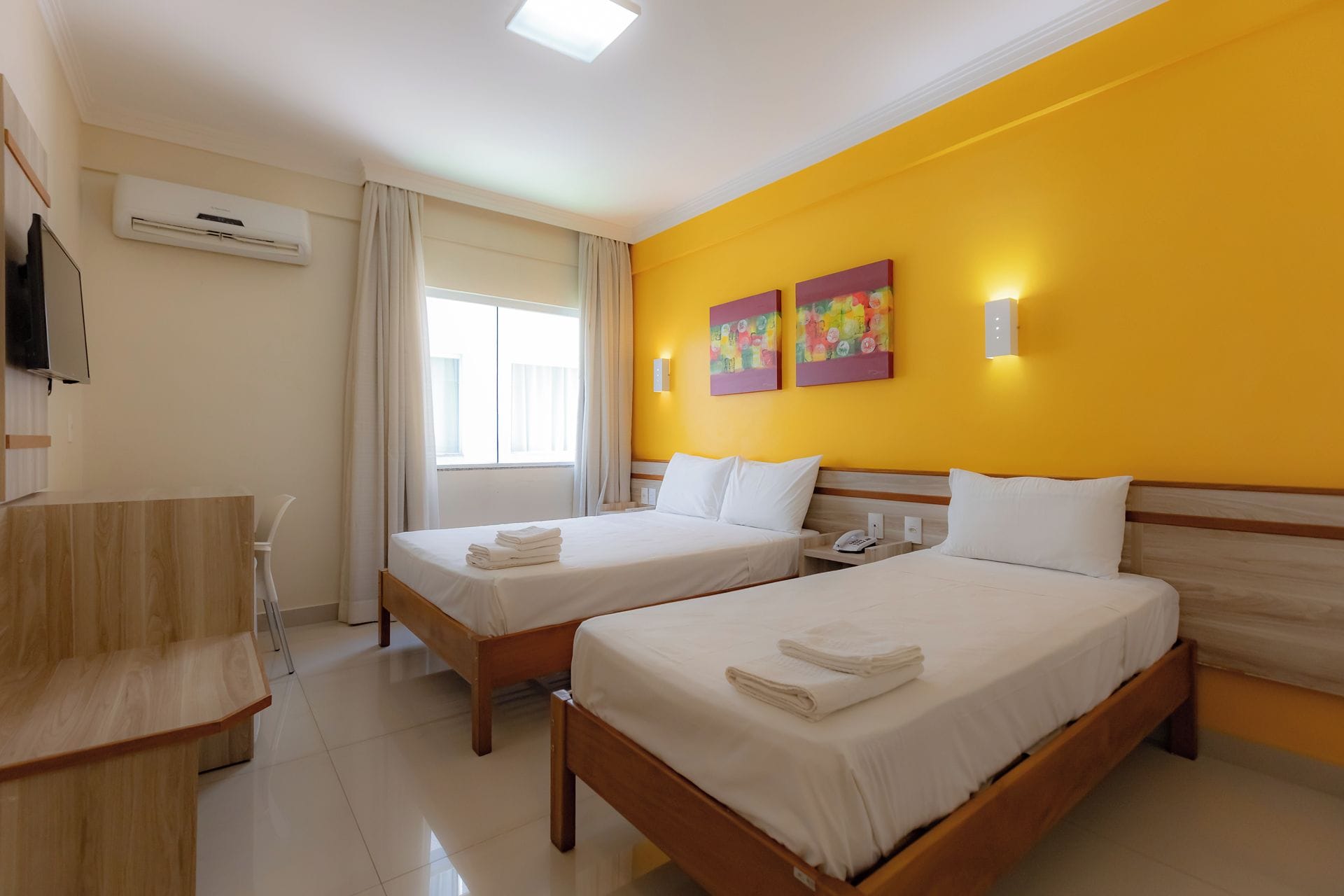 A hotel room with two beds, a desk, a TV, and a window with curtains.