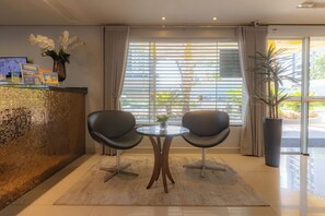 A reception area with a reception desk, two chairs, a small table, and a large window with blinds.