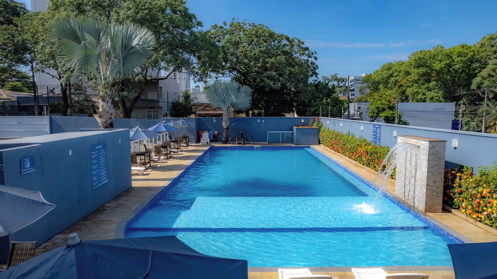 Una piscina con sillas de descanso y sombrillas, rodeada de árboles y un muro azul.