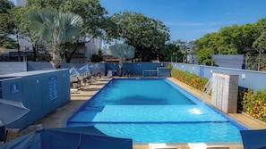 A swimming pool with lounge chairs and umbrellas, surrounded by trees and a blue wall. - Foz By Candeias (Foz do Iguaçu)