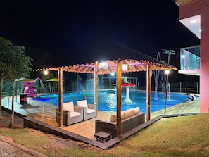 Un área de piscina al aire libre con una zona de estar techada, una fuente de agua y un edificio al fondo.