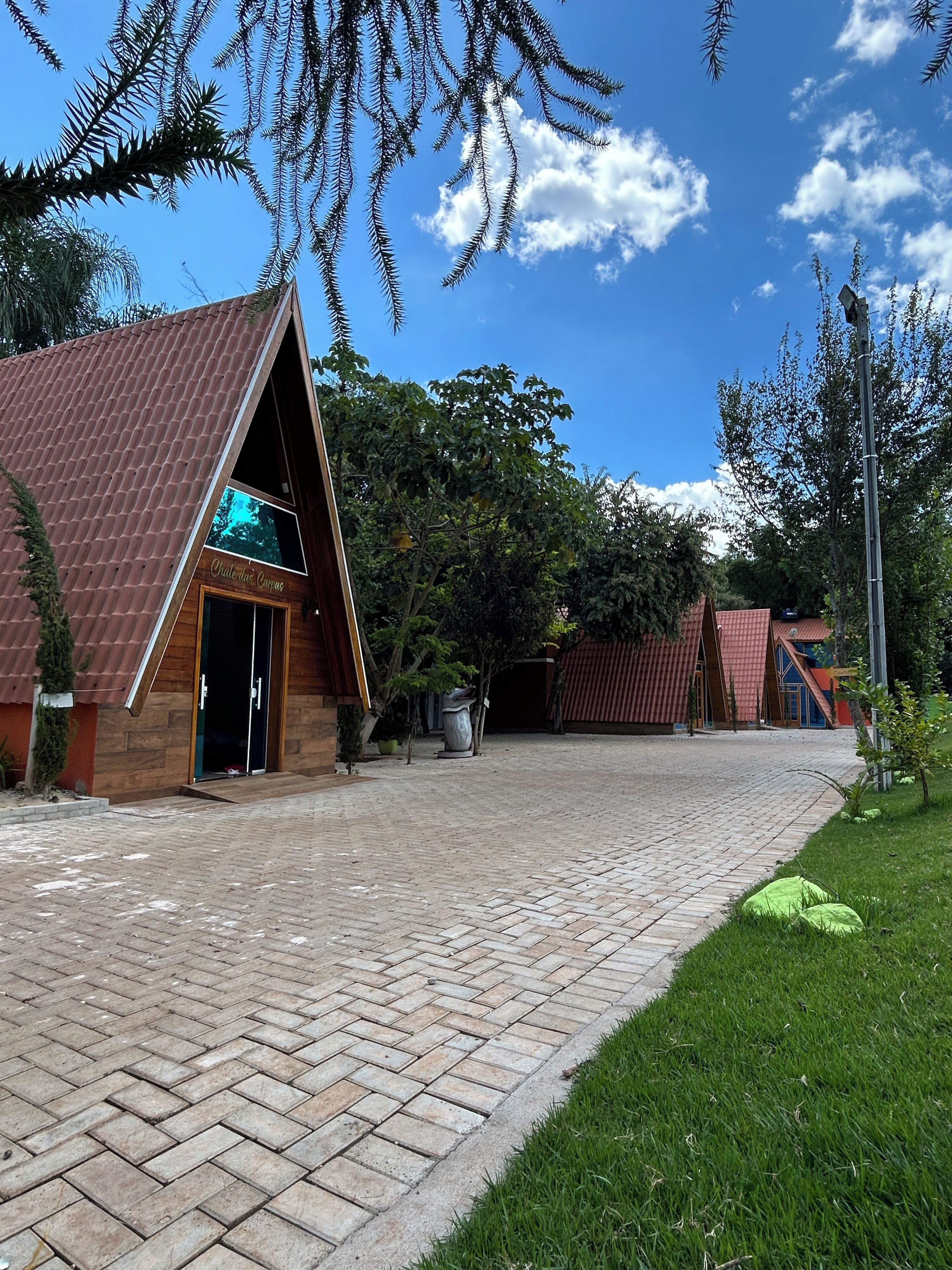 A building with a triangular roof and a wooden facade, a paved walkway, and surrounding greenery.