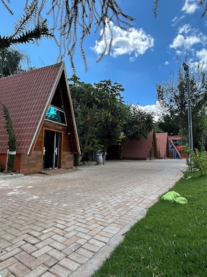 A building with a triangular roof and a wooden facade, a paved walkway, and surrounding greenery.