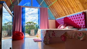 Un dormitorio con una cama grande, un sillón de felpa rojo y vistas al cielo a través de un ventanal.