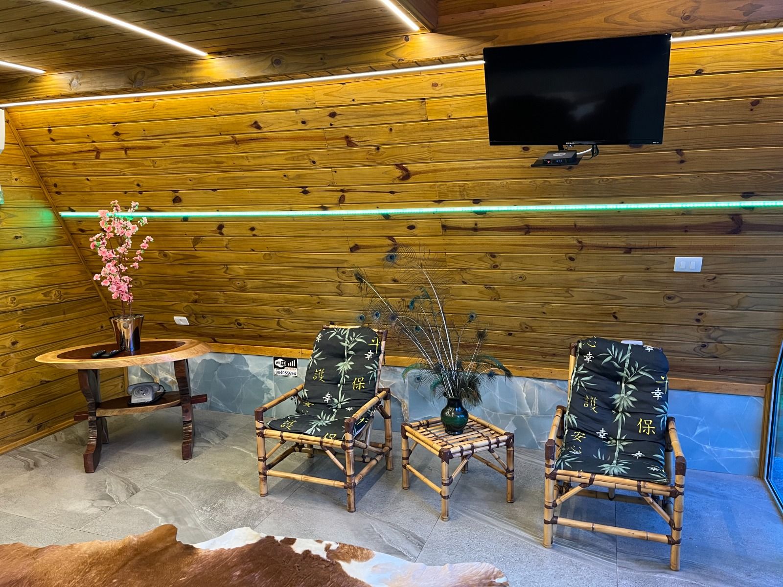 Una habitación con paredes de madera, un televisor de pantalla plana sobre una mesa, dos sillas de bambú, una pequeña mesa con un florero y un tapiz de cuero de vaca en el suelo.