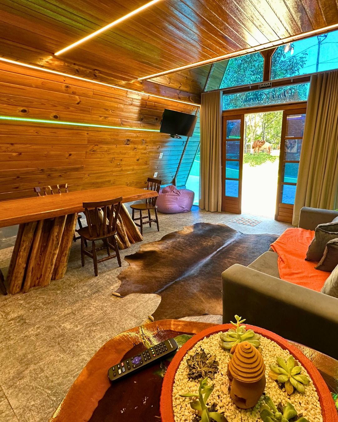 Una habitación con paredes de madera, un ventanal grande, un televisor de pantalla plana, una mesa de comedor con sillas, un sofá y un control remoto sobre una mesa.