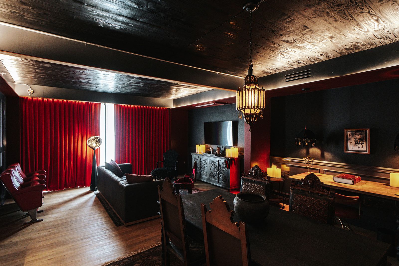 A dimly lit room with a red curtain, a leather sofa, a dining table with candles, and a vintage chandelier.