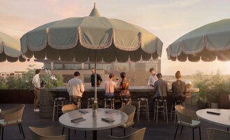 A rooftop bar with round tables and chairs, umbrellas, and a bar area with people.