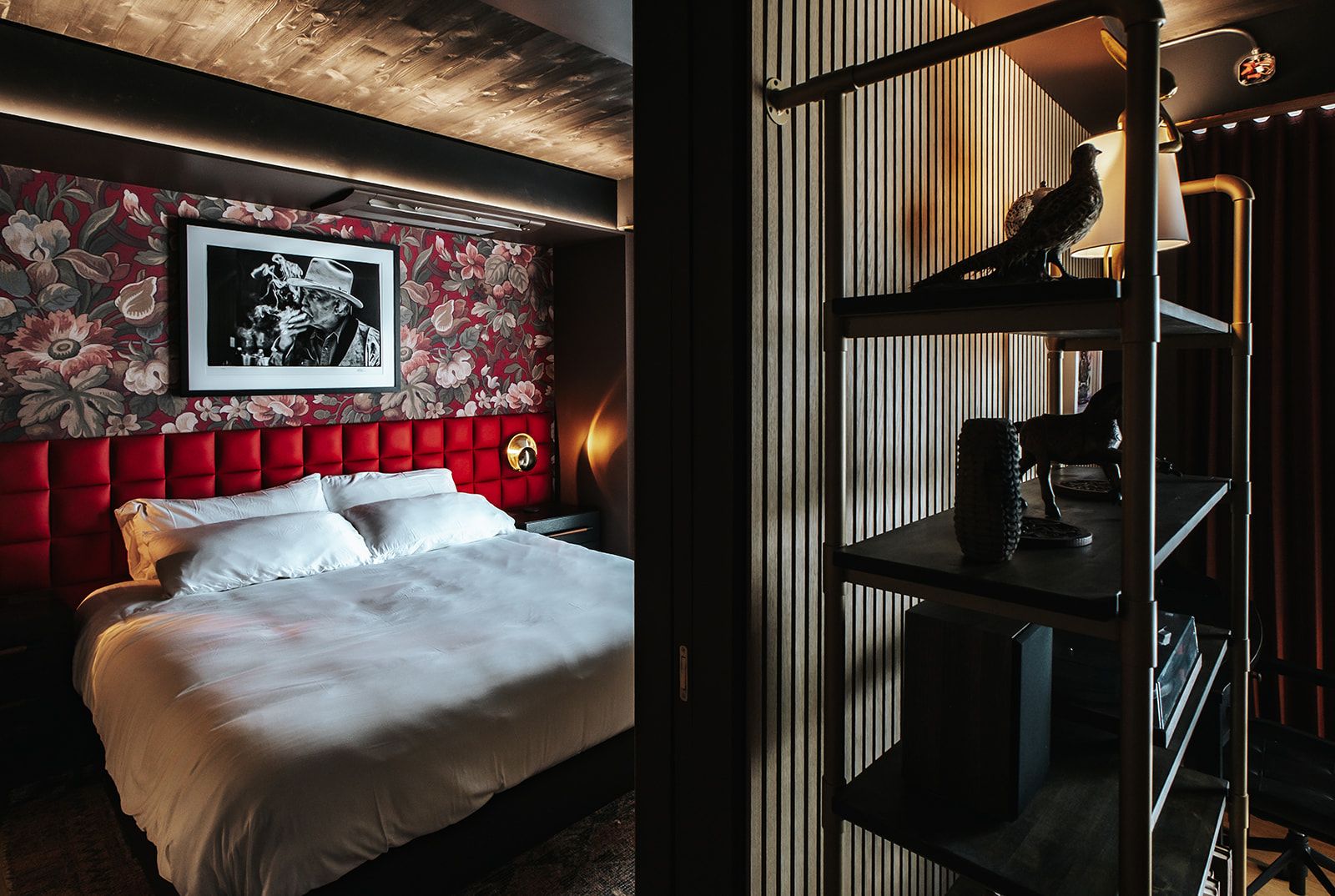 A hotel room with a red upholstered headboard, a framed black and white photograph, and a desk with a lamp.