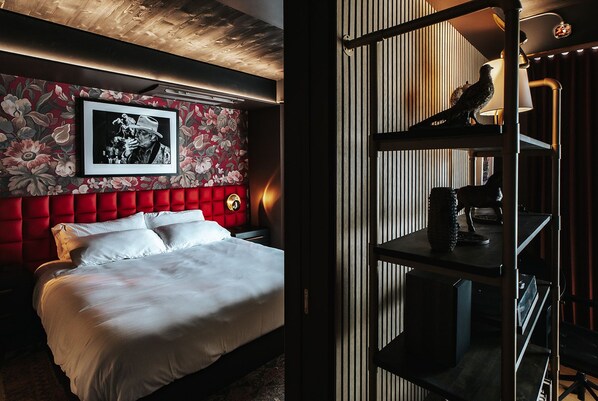 Une chambre d’hôtel avec une tête de lit recouverte d’un tissu rouge, une photographie en noir et blanc encadrée et un bureau avec une lampe.