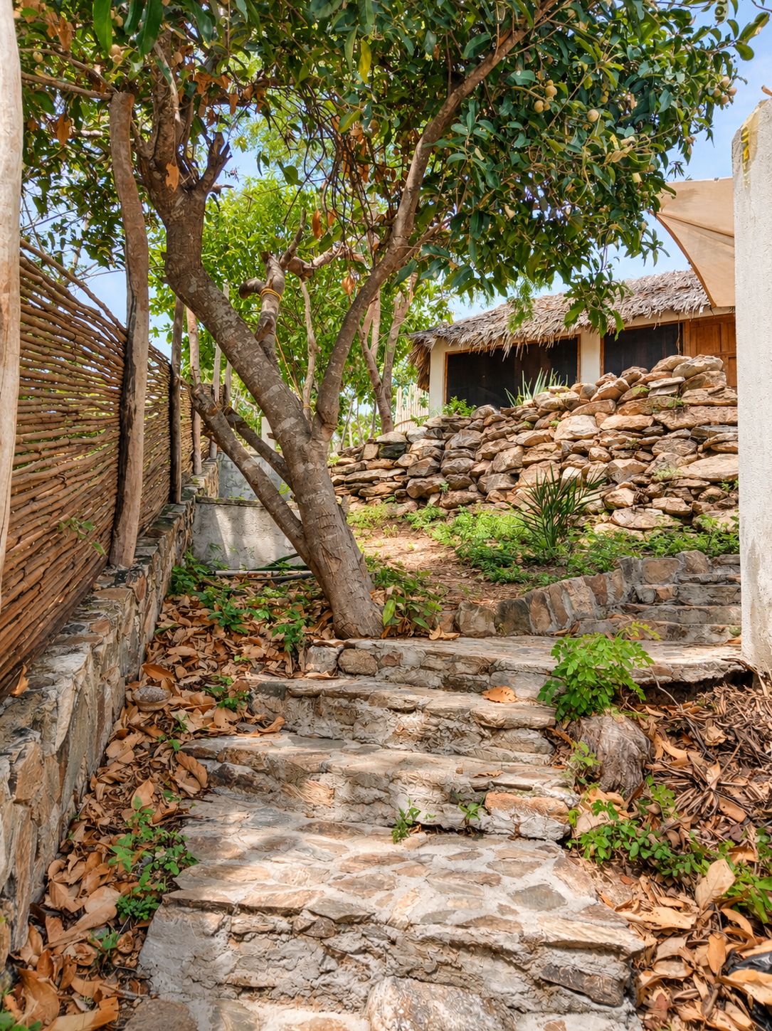 Una escalera de piedra que sube hacia un edificio con techo de paja, rodeado de árboles y un muro de piedra.
