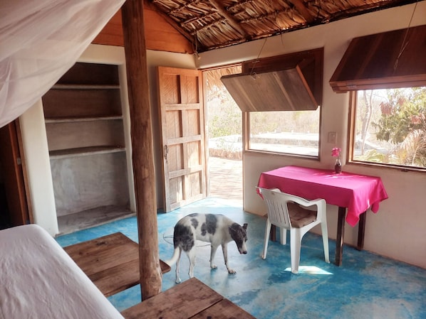 A room with a wooden bed, a table with a pink tablecloth, a dog, and a window with a view of greenery.