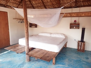 A single bed with a wooden frame and a white mattress, a mosquito net above, a small wooden cabinet with a cup, and a heater on a wooden stand.