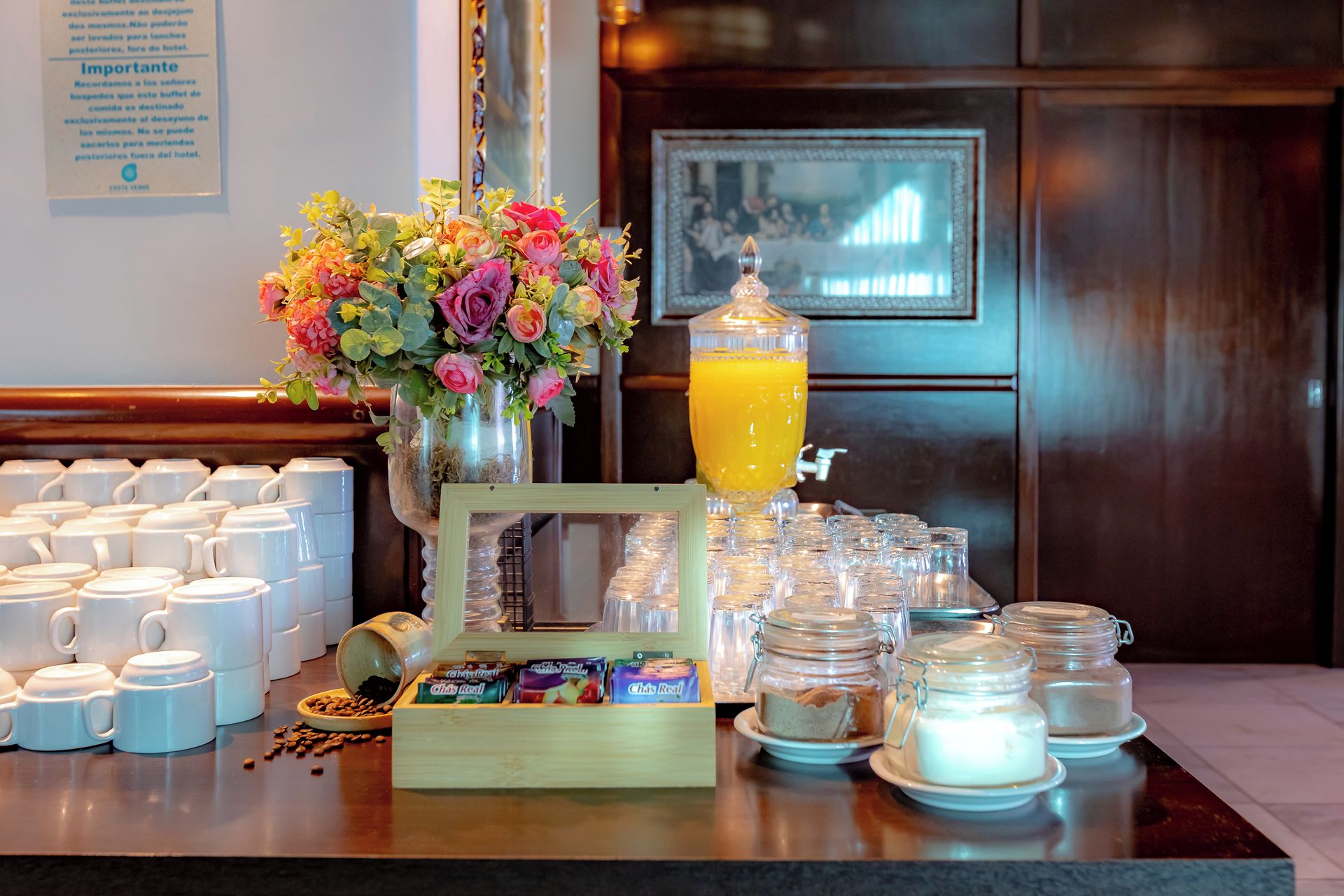 Montaje de un bufé de desayuno con tazas de café, un bol de frutas, un arreglo floral y un dispensador de jugo.