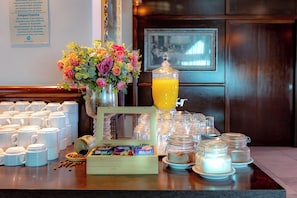 A breakfast buffet setup with coffee mugs, a fruit bowl, a flower arrangement, and a juice dispenser.