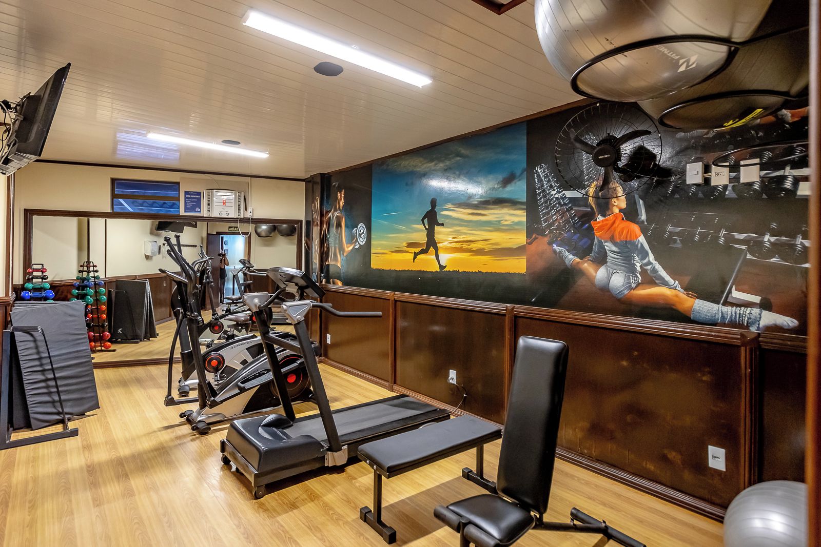 Un gimnasio con equipamiento de ejercicio, un mural y piso de madera.