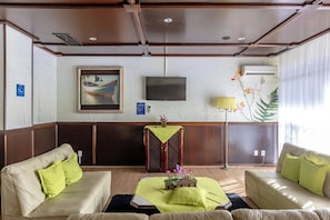 A hotel lobby with a seating area, a flat-screen TV, a decorative plant, and a wall-mounted air conditioning unit.