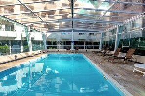 A swimming pool with a glass ceiling, lounge chairs, and a view of buildings. - Costa Verde By Candeias (Bombinhas)
