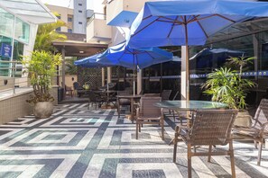 Un área de estar al aire libre con mesas y sillas, sombrillas azules y plantas en macetas.