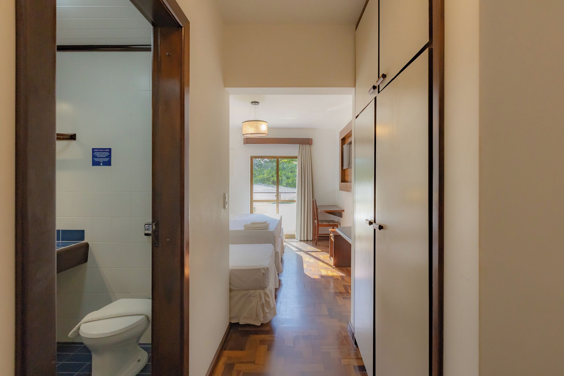 A hotel corridor with a wooden floor, a bed, a desk, and a bathroom with a toilet.