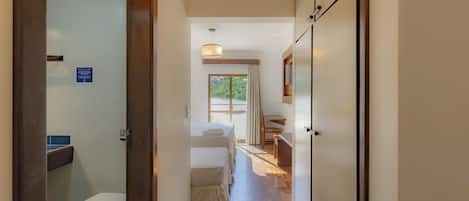 A hotel corridor with a wooden floor, a bed, a desk, and a bathroom with a toilet.