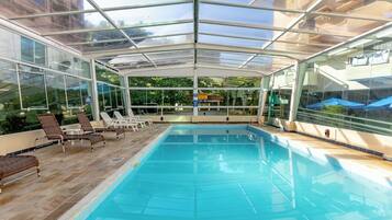 A clear glass-enclosed swimming pool with lounge chairs and a view of the outdoors.