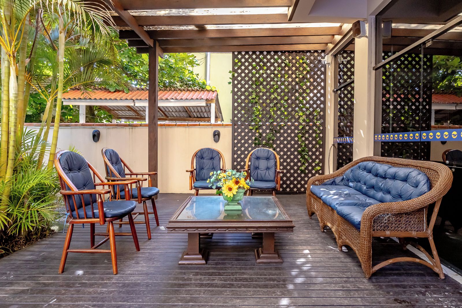 Un área de estar al aire libre techada con sillas de madera y un sofá de mimbre, una mesa con superficie de vidrio y un florero, y un muro de celosía con plantas trepadoras.