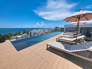 Ein Dachpool im Infinity-Design mit Liegestühlen und Sonnenschirmen mit Blick auf eine Küstenstadt.