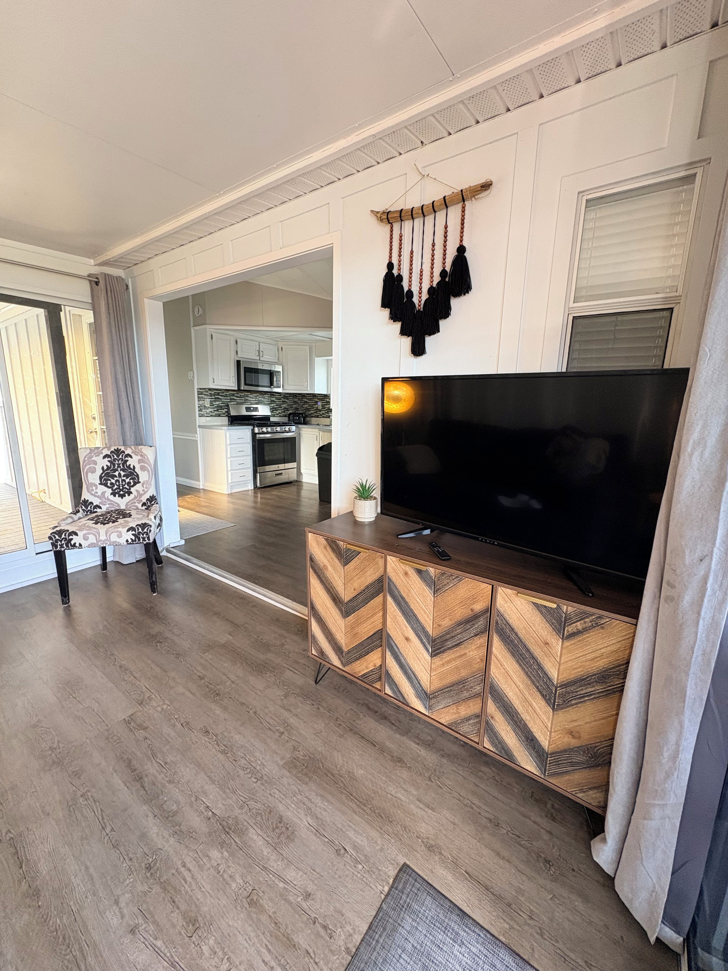 Un salon avec un grand téléviseur à écran plat posé sur un meuble en bois, un fauteuil à motifs et une vue sur la cuisine par une porte ouverte.