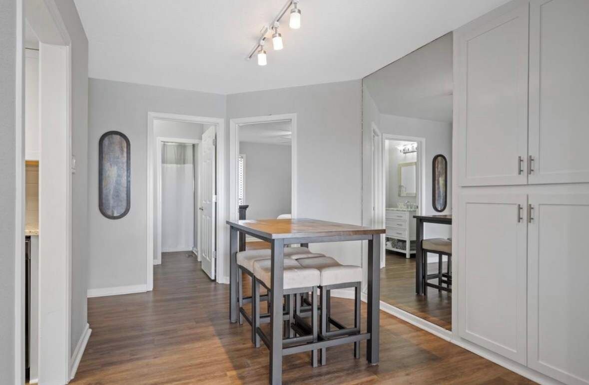 A modern kitchen with a wooden table and bar stools, a large mirror, and white cabinetry.