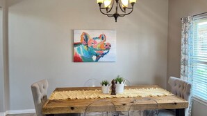 Un comedor con una mesa de madera, sillas transparentes, una araña y un cuadro de colores en la pared.