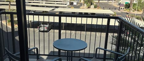 Un balcón con mesa y sillas, con vista a un estacionamiento y edificios a la distancia.