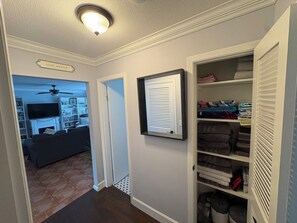 Ein Flur mit geschlossener Tür, einem gerahmten Schild, einem Kleiderschrank mit gefalteter Kleidung und einem Wohnzimmer mit Fernseher und Büchern.