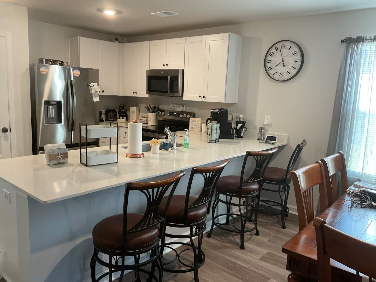 A modern kitchen with white cabinets, a stainless steel refrigerator, a microwave, and a coffee station. There are four bar stools and a dining table with chairs.