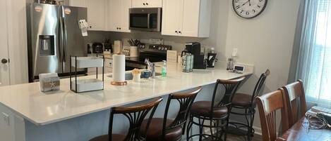 A modern kitchen with white cabinets, a stainless steel refrigerator, a microwave, and a coffee station. There are four bar stools and a dining table with chairs.