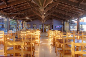 A spacious indoor area with rows of wooden tables and chairs, a kitchen area in the background, and a ceiling with exposed beams.