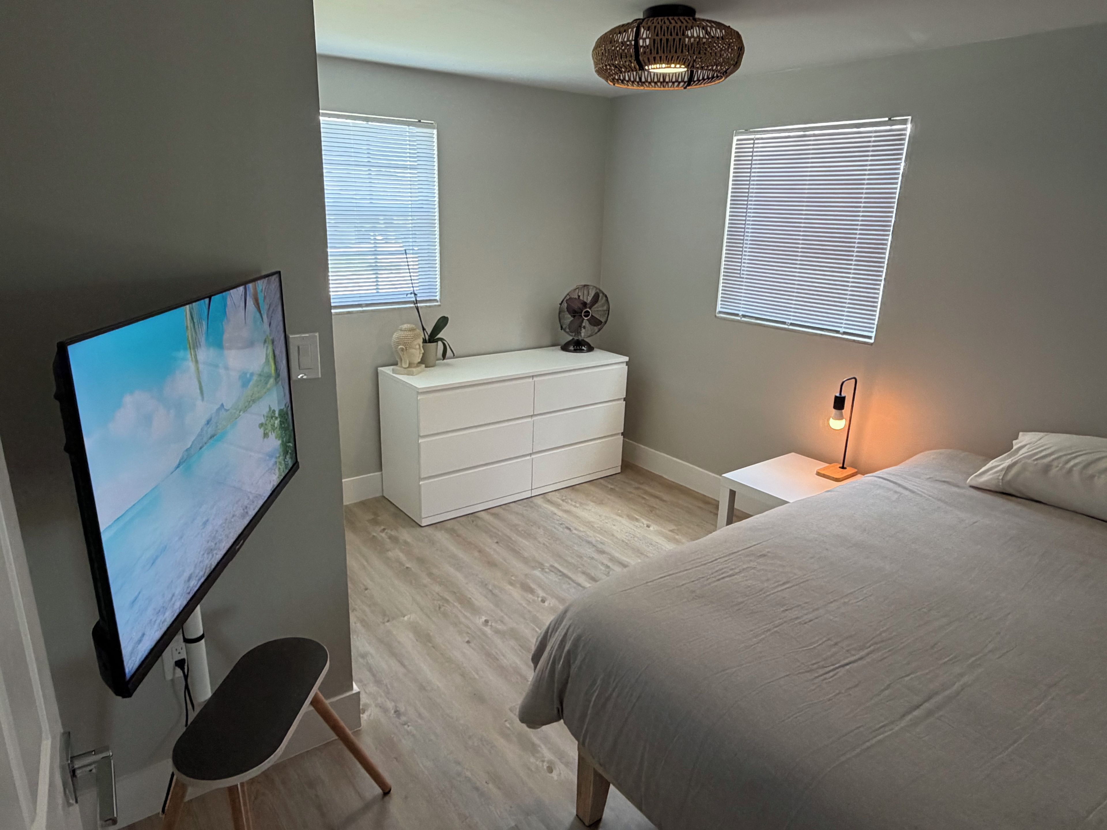 A bedroom with a bed, a dresser, a TV, and two windows with blinds.