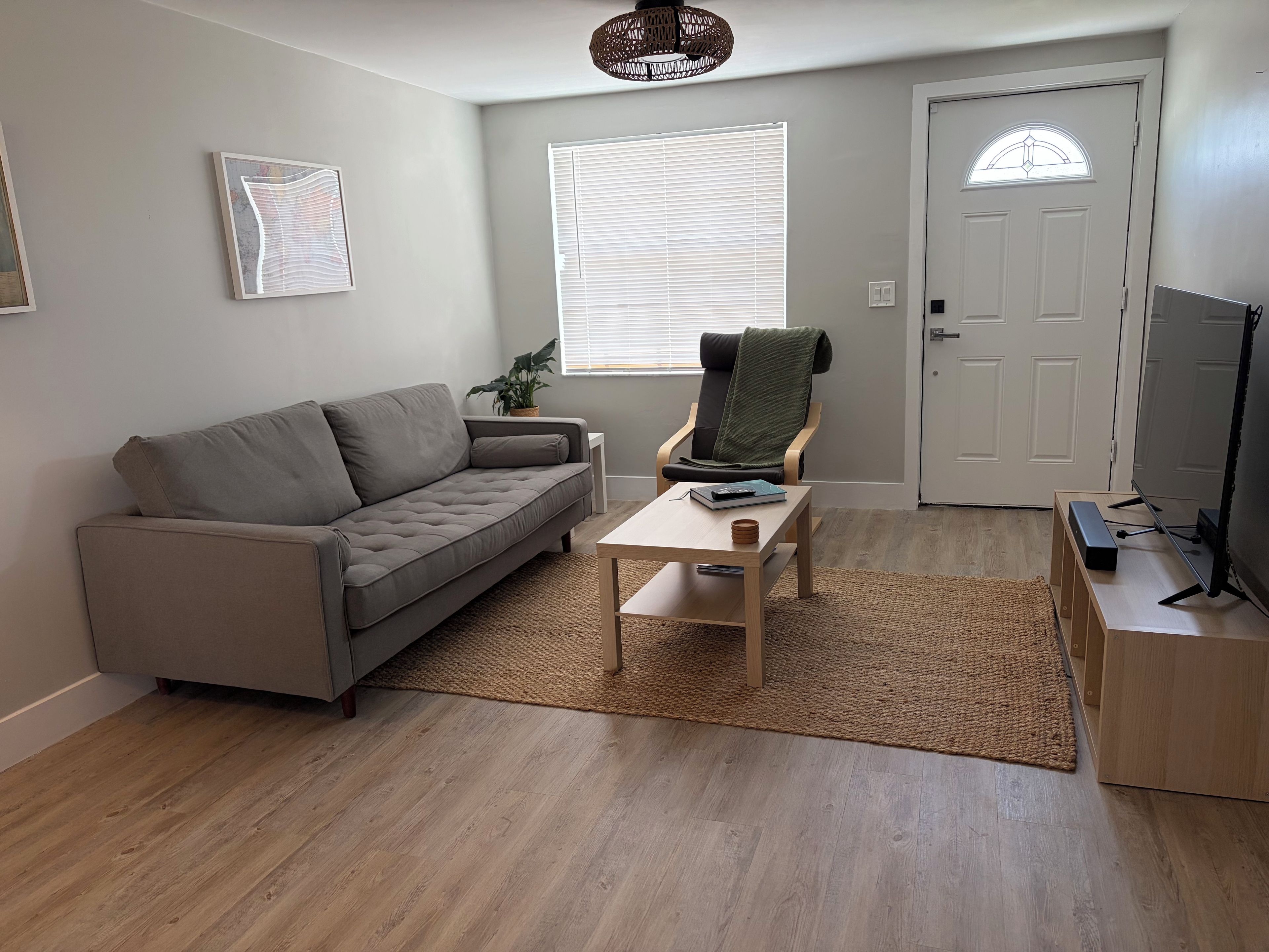 A living room with a grey sofa, a small table with a remote control, a chair with a blanket, a television, and a door with a window.