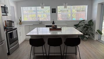 Een moderne keuken met witte kastjes, een centraal eiland met barkrukken, een inbouwoven en een groot raam met zonwering.