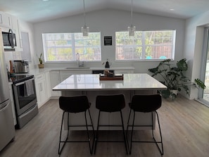 A modern kitchen with white cabinetry, a central island with bar stools, a built-in oven, and a large window with blinds.