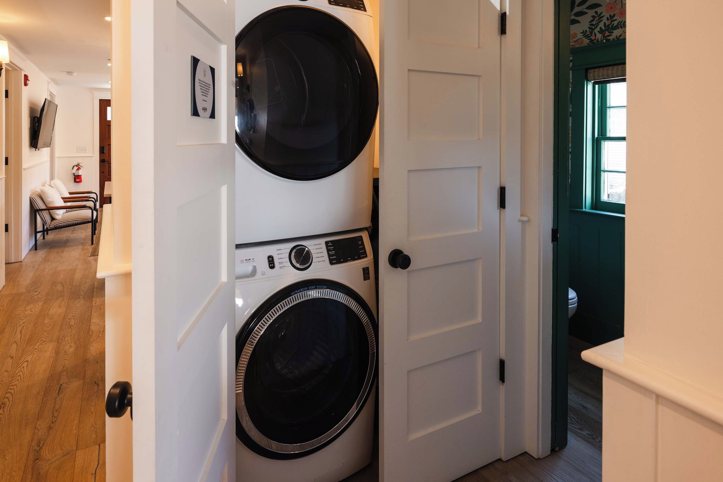 A laundry room with a front-loading washer and dryer, a door with a handle, and a hallway leading to a seating area with a fire extinguisher.