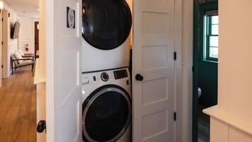 A laundry room with a front-loading washer and dryer, a door with a handle, and a hallway leading to a seating area with a fire extinguisher.