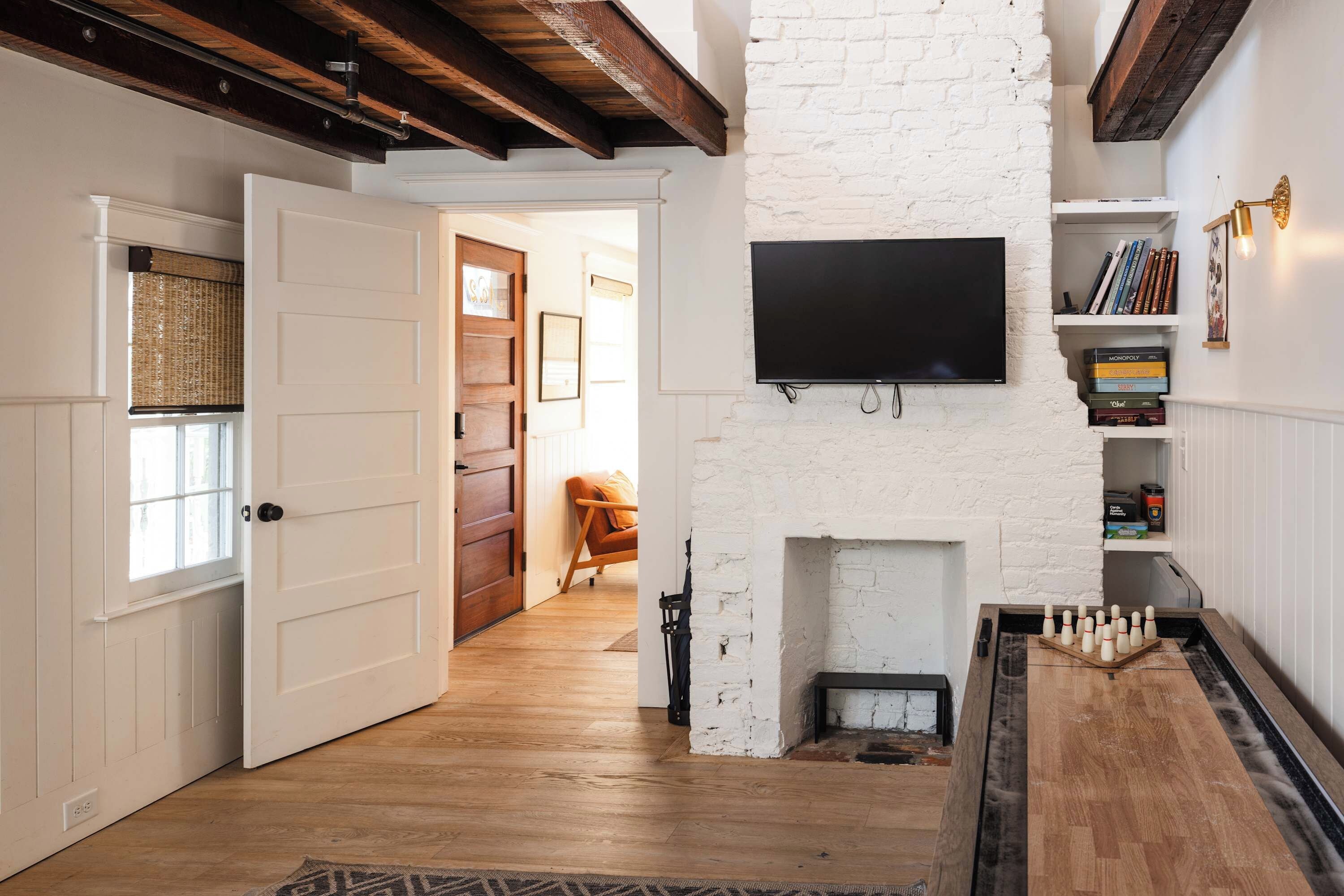 A room with a white brick wall, a flat-screen TV mounted above a fireplace, a wooden floor, and a door leading to another room.