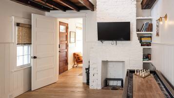 A room with a white brick wall, a flat-screen TV mounted above a fireplace, a wooden floor, and a door leading to another room.