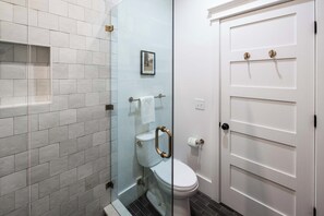 A bathroom with a toilet, a glass shower enclosure, and a white door with a gold handle. - Downtown 3bd Retreat, Newly Updated & Bright! (Charleston)